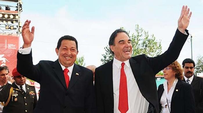 Hugo Chávez y Oliver Stone juntos con una mano en alto saludando. Al fondo otras personas y Nicolás Maduro.