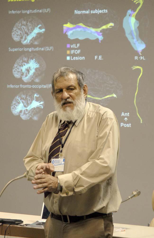 Hombre de barba blanca que viste camisa de mangas largas, se ven micrófonos y representaciones del cerebro humano en la parte posterior.