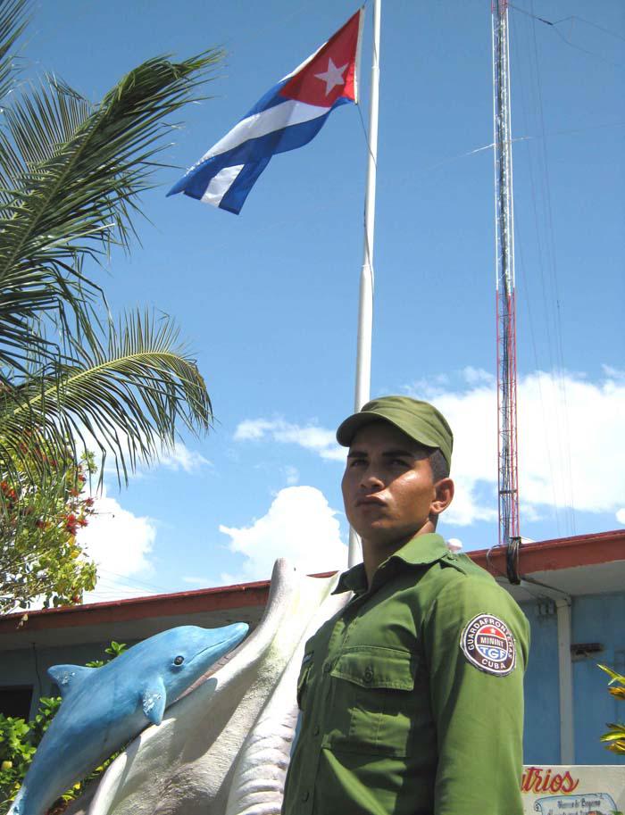 Guardafrontera y al lado una escultura de un delfín, en la parte trasera se ve una bandera que ondea a lo alto.