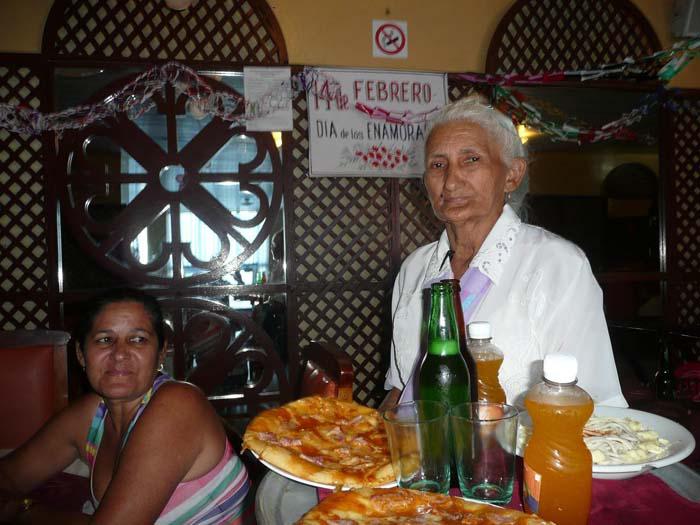 Señora que lleva unos platos con alimentos y dos pomos de refresco y dos botellas de cerveza. Una cliente sentada a un costado.