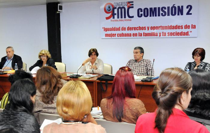 Varias mujeres sentadas escuchan a una que les habla desde una mesa que se encuentra frente a estas. En la mesa se encuentran dos mujeres más y dos hombres.