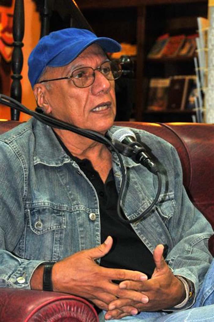 Víctor Casaus en chaqueta de mezclilla hablando por un micrófono.
