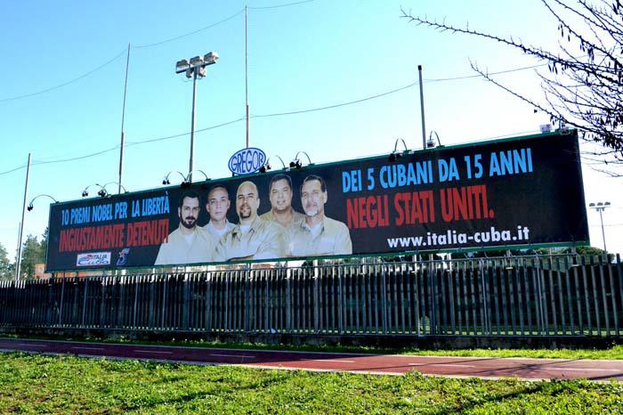 Valla en la cual hay una imagen de los cinco héroes cubanos y un letrero en azul rojo en italiano.