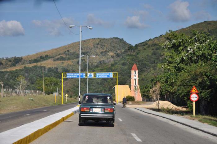 Un carro transita por una carretera, al fondo de la fondo se ven dos elevaciones montañosas.
