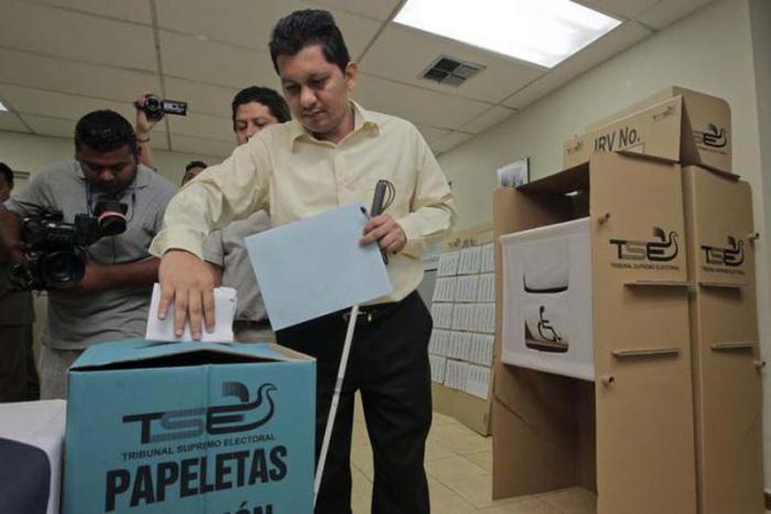 Un hombre deposita su voto en una urna  de cartón en la que se lee TSE PAPELETAS. A su lado un recinto para votar y detrás de él se ve otro hombre y un camarógrafo.