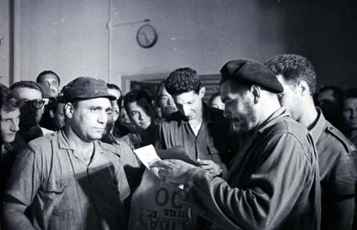 El Comandante Ernesto Che Guevara realiza una jornada de trabajo voluntario en los Molinos de Trigo José A. Echeverría, en Regla, y cose el saco 100 000. En el centro el Comandante Juan M. Castiñeiras y detrás del Che el capitán Jesús Suárez Gayol. 