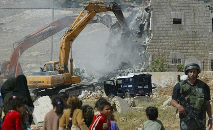 Under the watchful eye of an Israeli border police officer, right, Palestinians look on during a demolition by Israeli authorities of the house belonging to the Zabouah family, in the West Bank village of Anata near Jerusalem Wednesday, June 2, 2004. The authorities demolished the two-storey building that used to house about 20 people, saying the house had been built without the proper permits but Palestinians say Israel rarely issues them permits as part of a policy to keep Arabs out of the Jerusalem area. A total of about 80 homes have been demolished in east Jerusalem area since the  beginning of 2004, according to the Israeli Committee Against House Demolitions organization. (AP Photo/Oded Balilty)