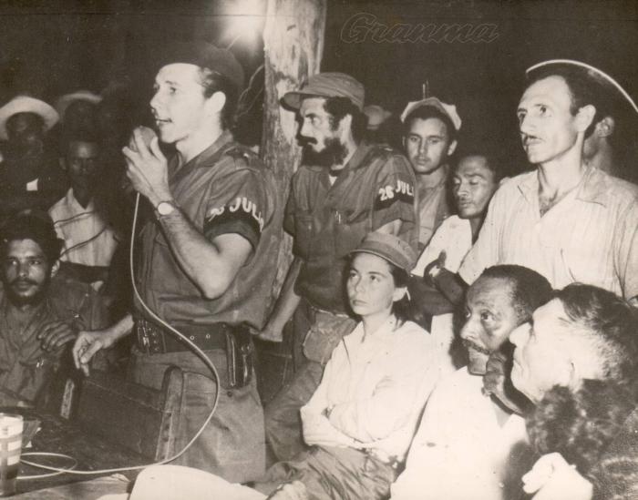 Primer congreso campesino en armas, celebrado el 21 de septiembre en Soledad de Mayarí, en territorio del II Frente guerrillero. En la foto Raúl Catro haciendo uso de la palabra, Vilma Espín sentada tras él y otros guerrilleros y campesinos (José Ramirez Cruz), APOYADO AL POSTE Jorge Serguera Riverí